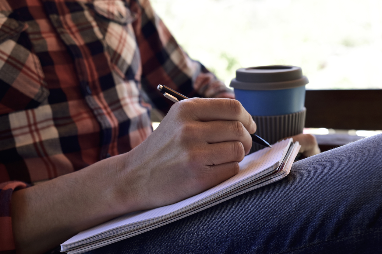closeup of a young caucasian man wearing jeans and a plaid shirt writing with a pen in a notebook in the porch of a house or a ranch