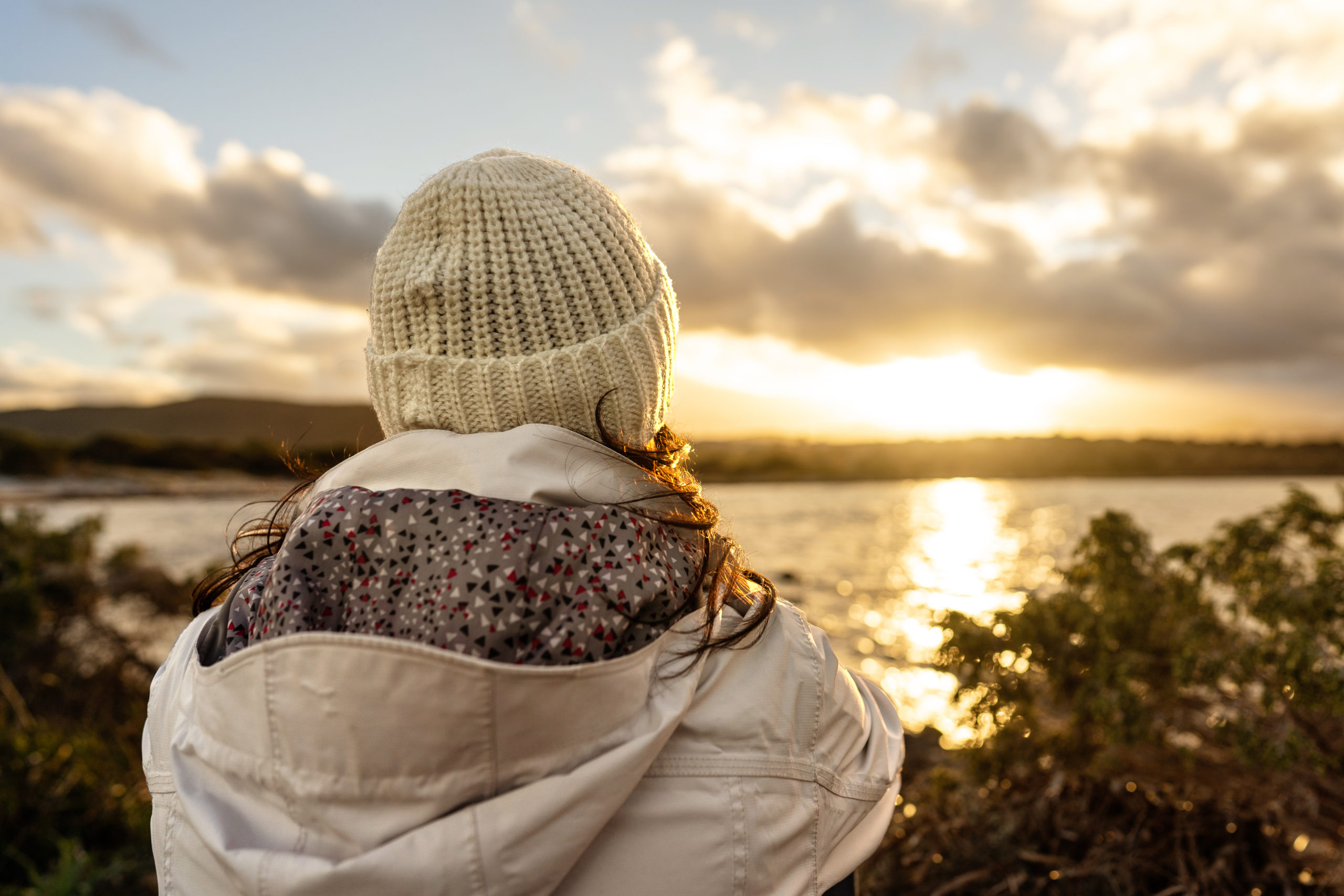 Pensive person view from back looking at the horizon. Unrecognizable woman seen from behind dressed in winter wool hooded clothes looks at a wonderful sunset or sunrise with clouds over the sea
