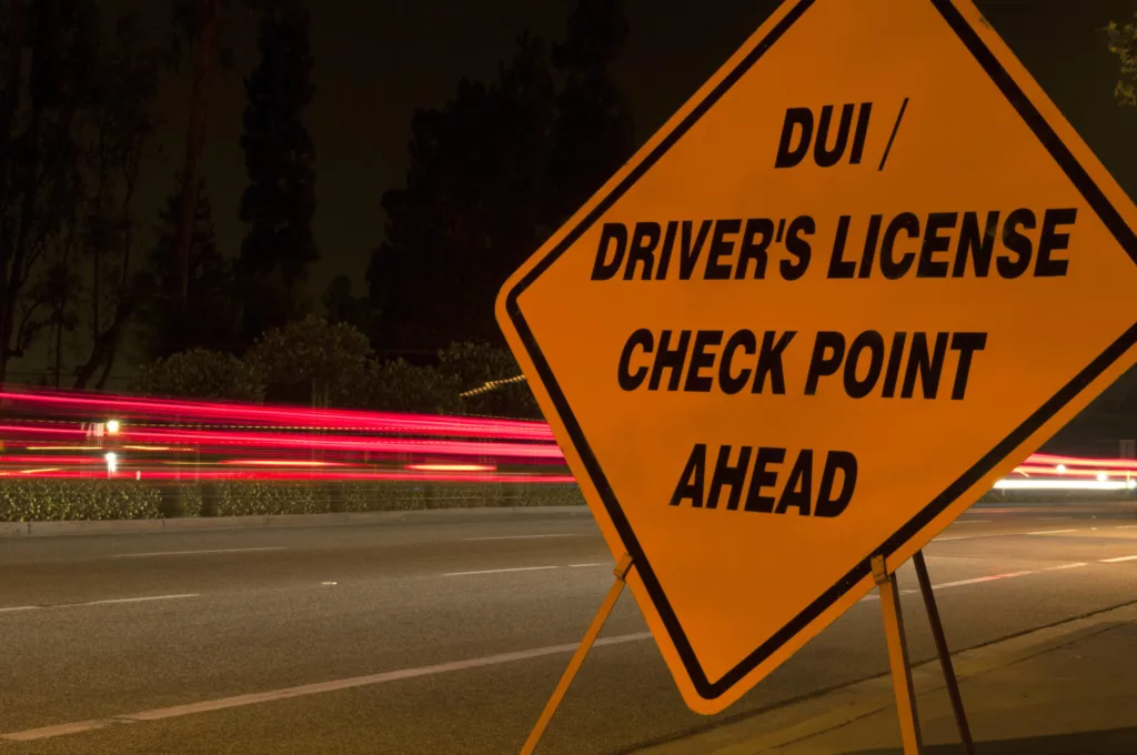 Orange DUI 'sobriety' and driver's license checkpoint ahead sign along side of road with car light trails in background